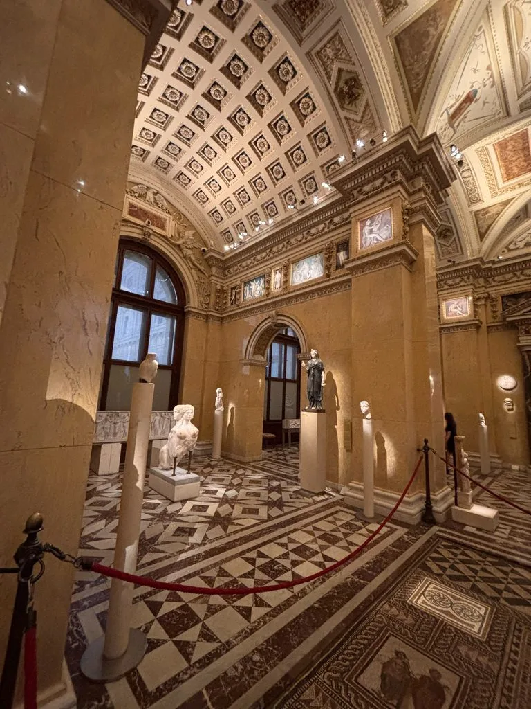 Museum gallery: Ornately decorated museum hall with marble floors, vaulted ceilings, framed artwork, and classical sculptures on display